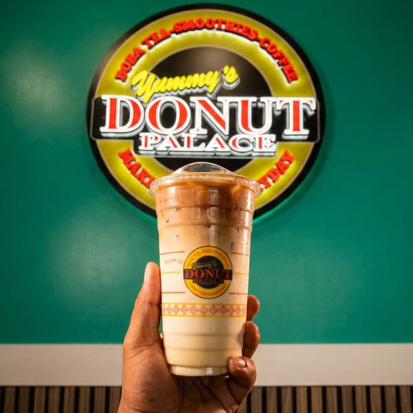 Hand holding a branded Donut Palace takeaway cup beneath a circular Donut Palace sign against a teal wall.