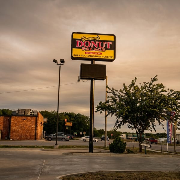 Yummy’s Donut Palace roadside sign stands above an empty parking lot, with trees, neighboring businesses, and a cloudy sunset sky.