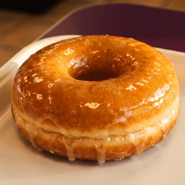 Glazed ring doughnut with icing dripping between its layers, resting on a white tray in a warmly lit restaurant.