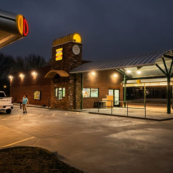 A warmly lit drive-through restaurant at dusk, with covered lanes, brick exterior, illuminated signs, and a parked Chevrolet pickup.